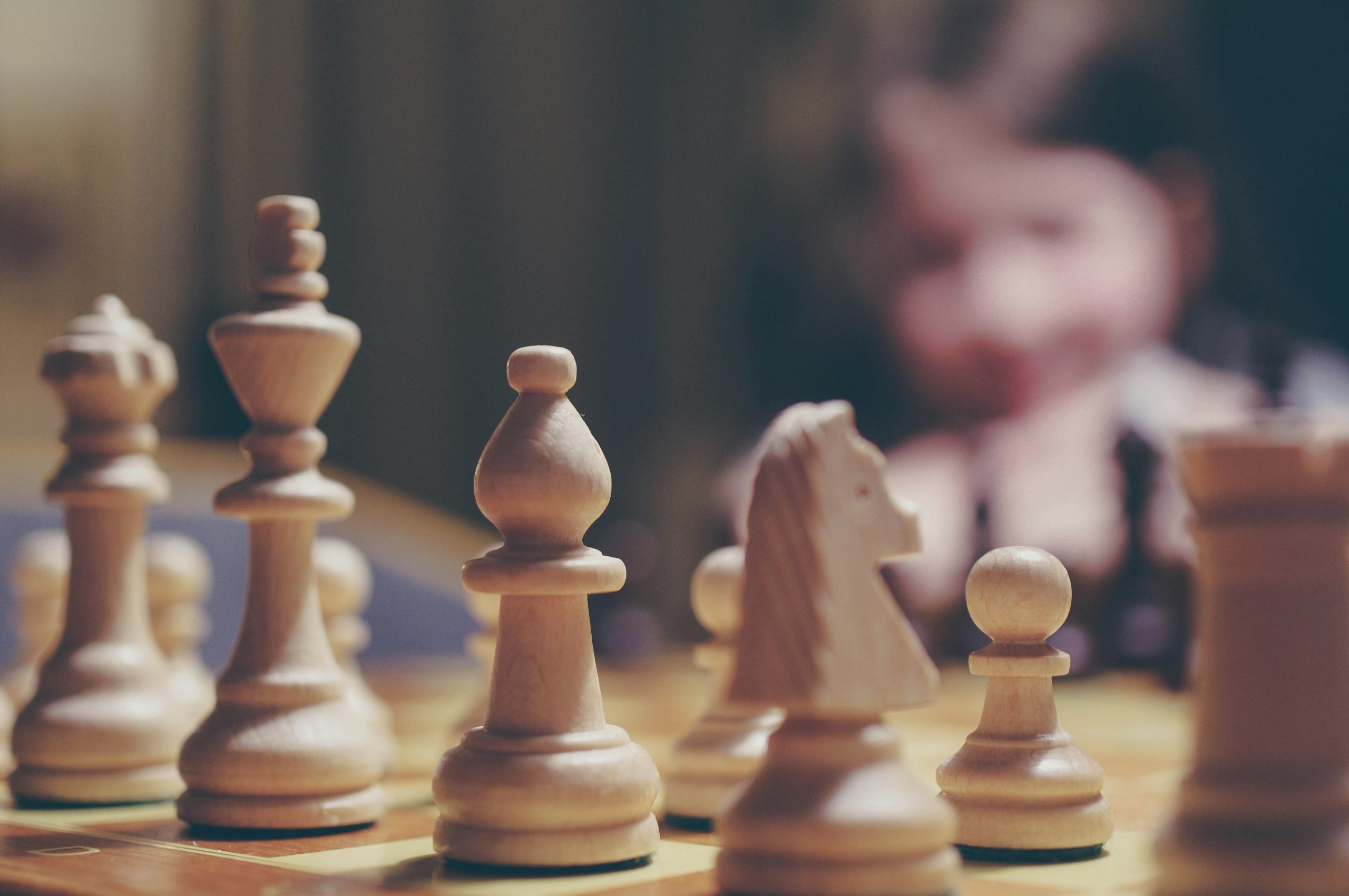 Photograph of wooden chess pieces on a chess board with a blurred person in the background