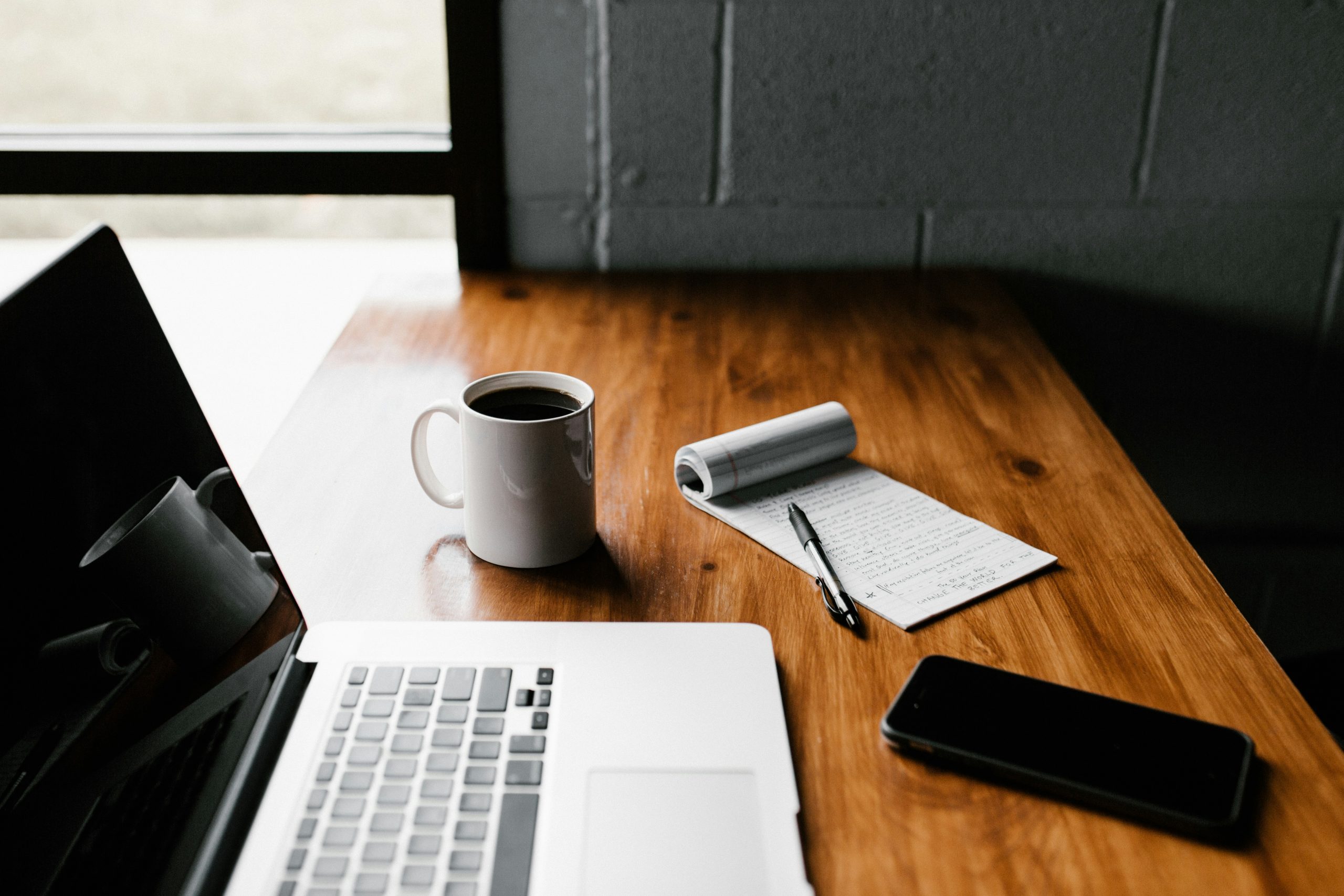 Photograph of a laptop, mouse, coffee cup, smartphone, and notepad on a wood desk