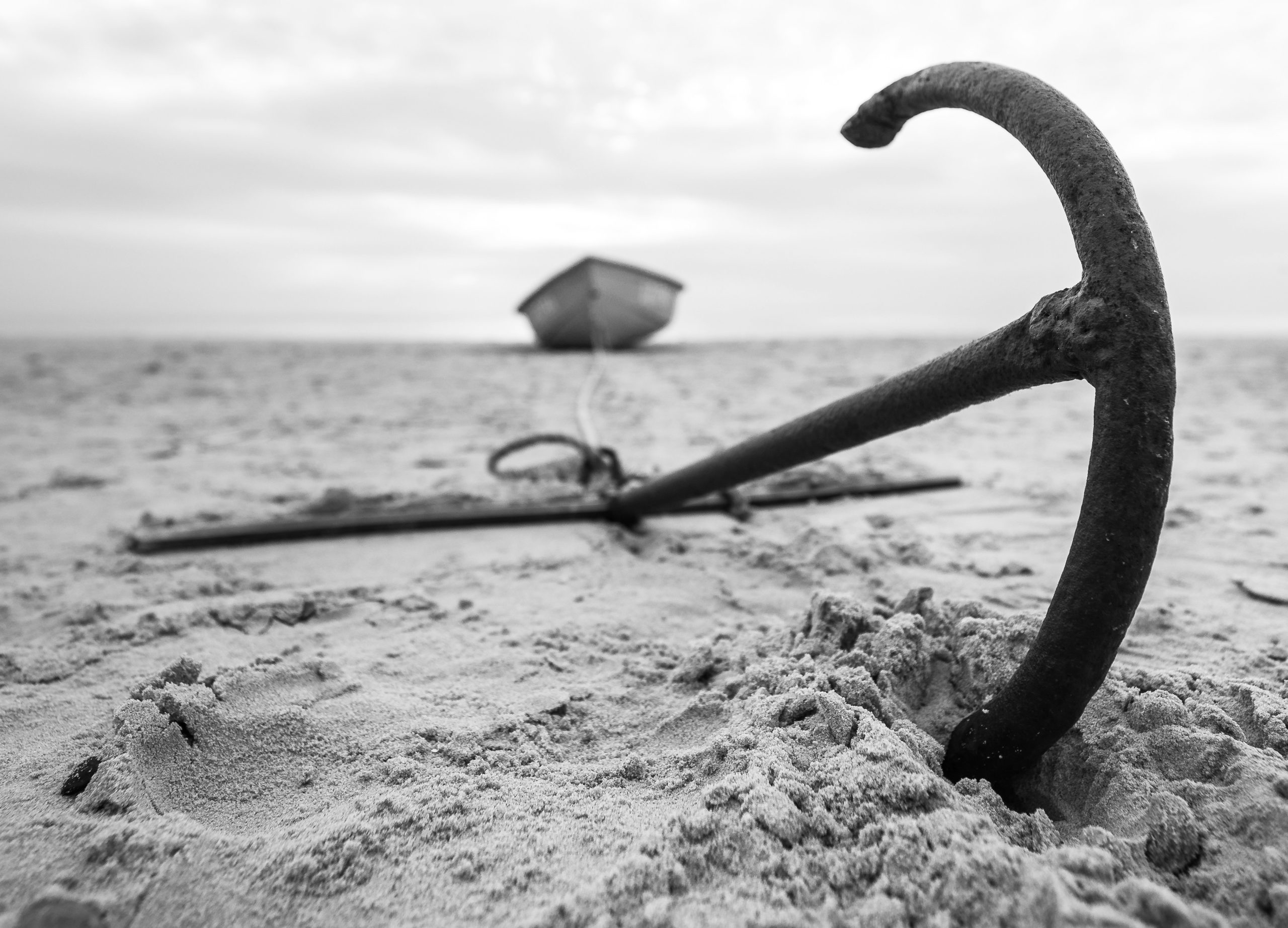 Black and white photograph of an anchor on a sandy beach with a small boat in the background