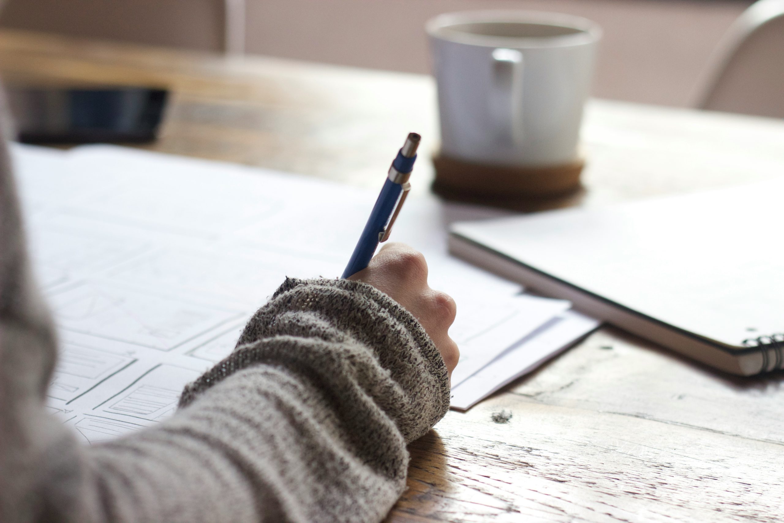 Photograph of a hand holding a pen while writing a blurred outline on paper with a mug in the background