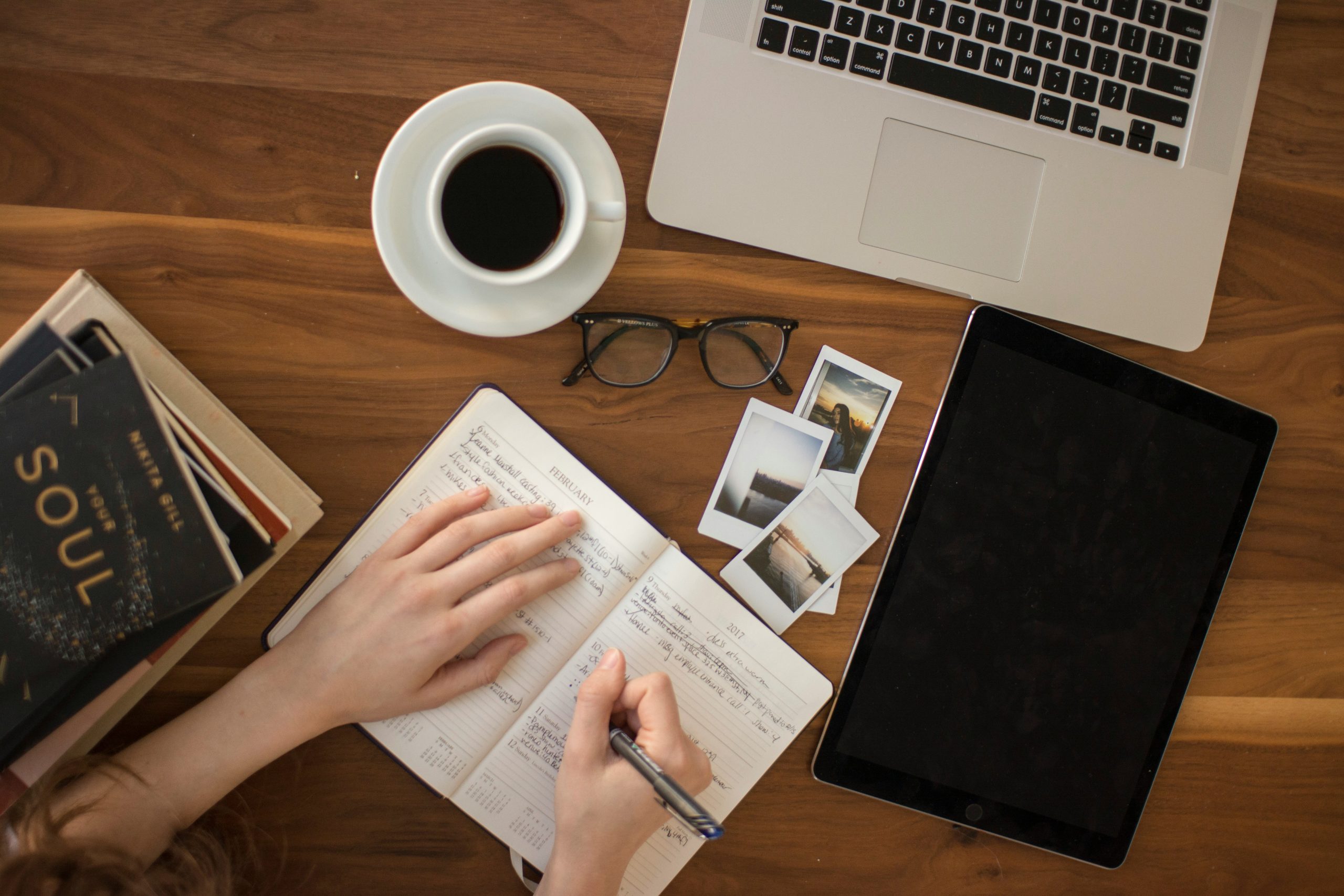 Photograph of a person at a wooden desk writing in a notebook with a laptop, photos, glasses, a cup of coffee, and a folder on it