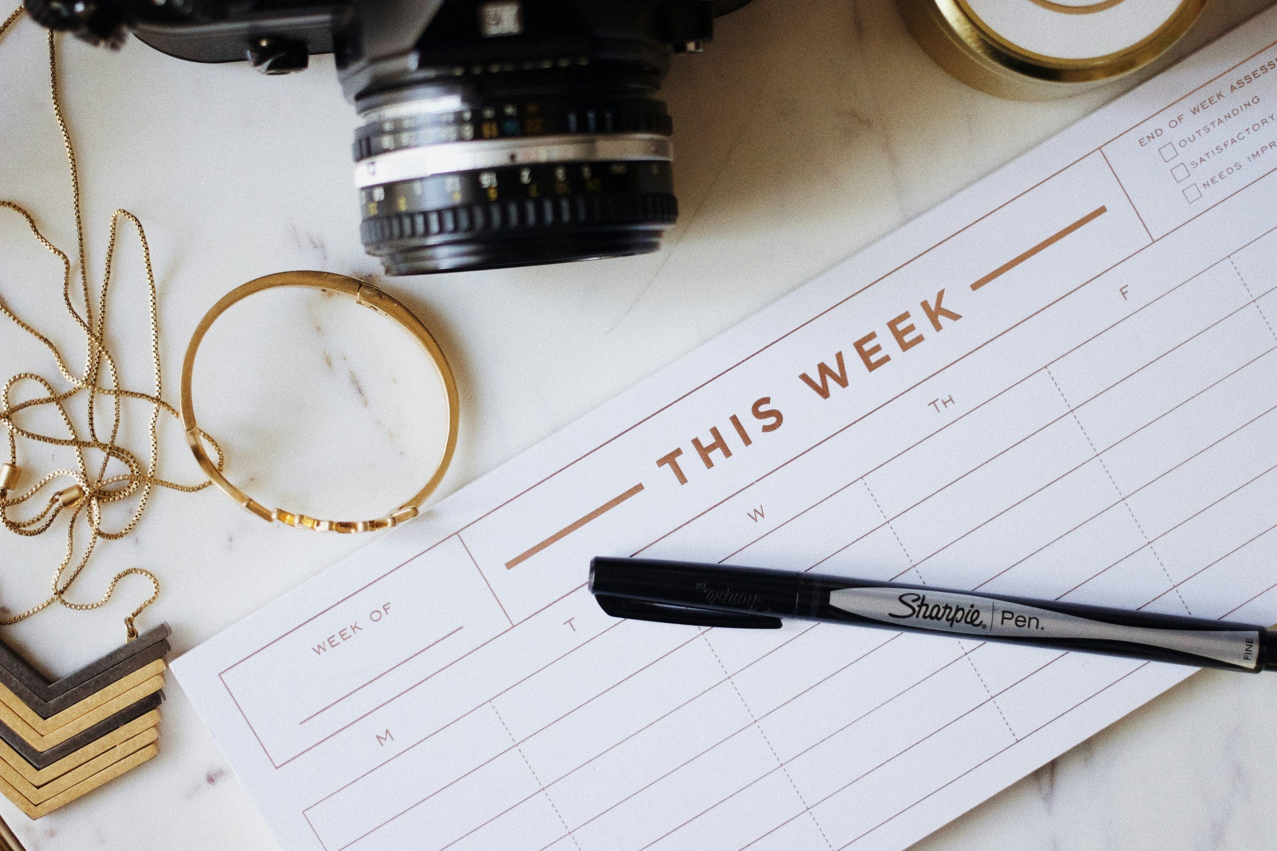 Photograph of a blank weekly planner on a desk with a camera, pen, and bracelet near it