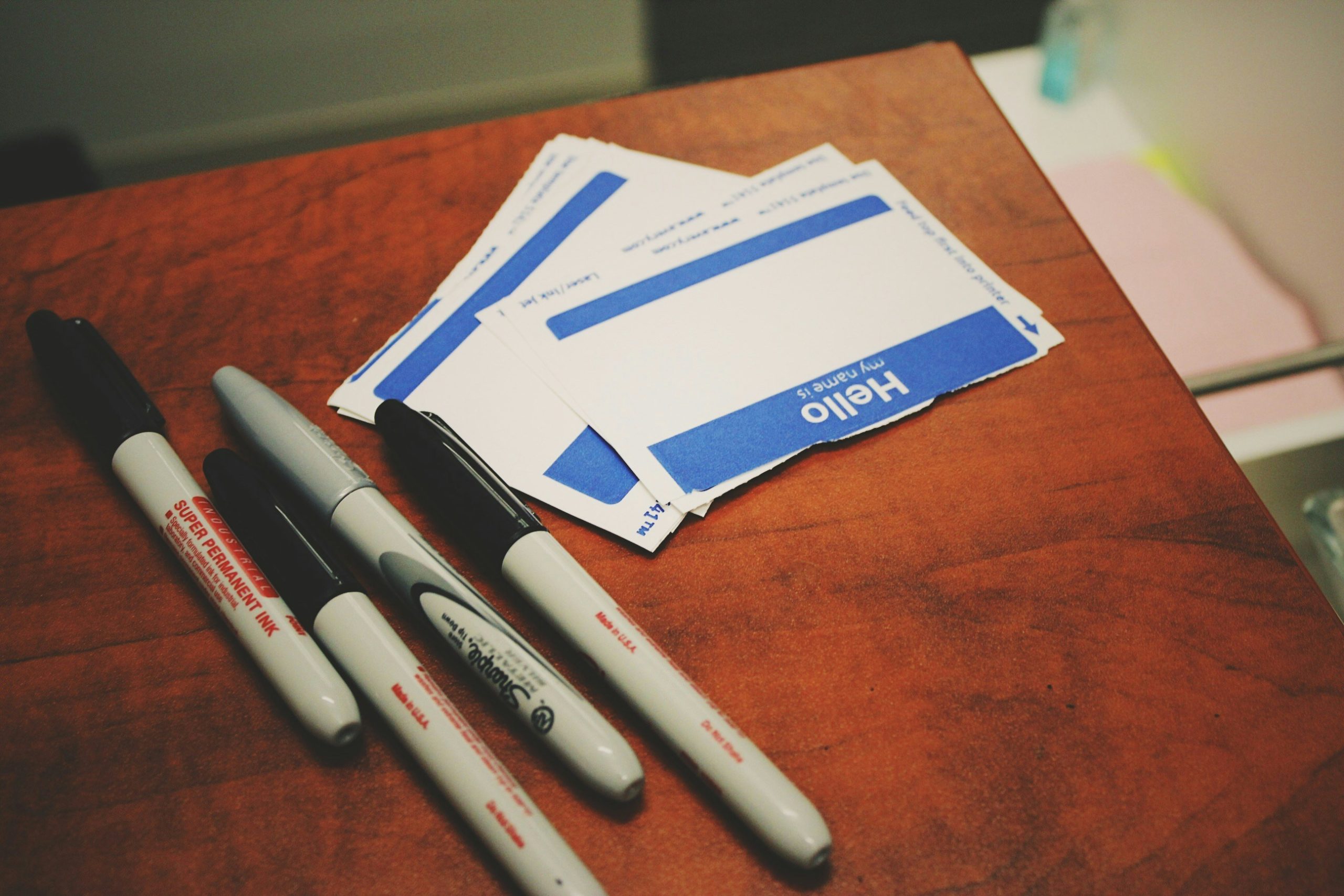 Photograph of blank name tags on a wooden desk next to four sharpies