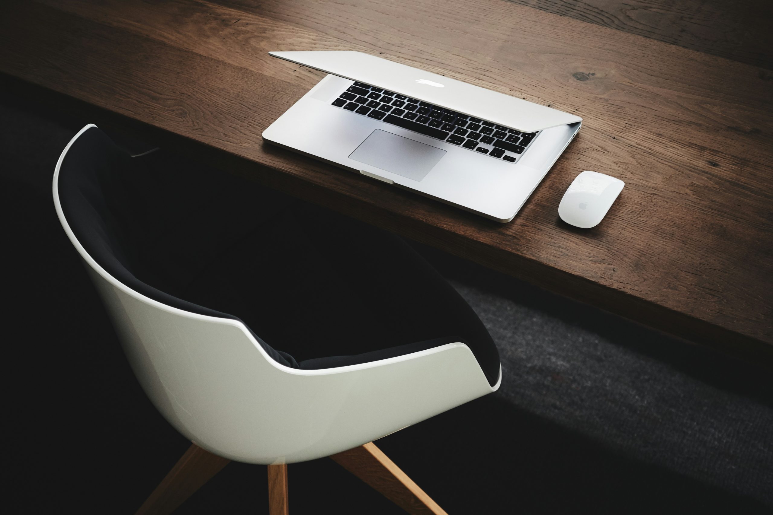 Photograph of a half closed macbook on a wooden desk with a mouse next to it and a white chair with wood legs in front